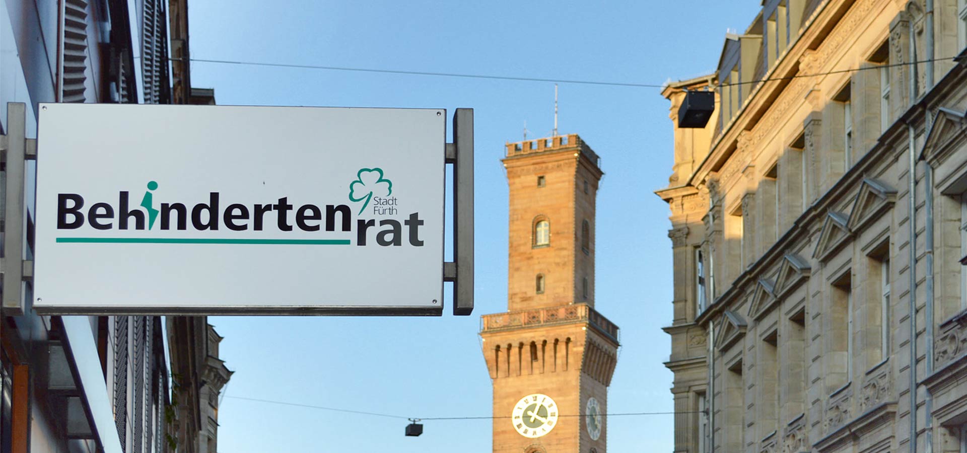 Ein Schild an einem Gebäude in Fürth mit dem Logo des Behindertenrats, im Hintergrund sieht man den Turm von Rathaus.